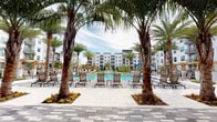 This outdoor amenity space is a standout resort-style pool deck designed to deliver a vacation-like experience within a contemporary residential community. At its center sits a large, crystal-clear swimming pool framed by towering palm trees that lend a tropical ambiance to the surroundings. The expansive pool deck is finished in elegant patterned stone pavers in complementary shades of gray, providing a clean and modern aesthetic throughout. Generous rows of sling-style lounge chairs are arranged across the deck, offering ample space for sunbathing and relaxation. Colorful seasonal flower beds punctuate the landscaping around the palm tree bases, adding vibrant accents to the otherwise refined palette. The perimeter of the space is flanked by the multi-story residential buildings, creating an enclosed courtyard feel that enhances privacy and atmosphere. Covered seating areas with ceiling fans and sleek outdoor furniture are positioned adjacent to glass-fronted interior amenity spaces, seamlessly connecting the outdoors to indoor facilities such as a fitness center. Decorative planters with lush tropical foliage are thoughtfully placed throughout the hardscape, softening the architectural lines. The entire space is open to the sky, flooding the deck with abundant natural light and reinforcing its inviting, sun-drenched character.