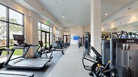 Located in Apex, NC, this fitness center offers a generously sized, single-floor layout divided into two dedicated exercise rooms. The main workout area is an expansive, open space defined by soaring ceilings, ceiling fans, and dramatic floor-to-ceiling windows along an entire wall, flooding the room with natural light and providing views of the surrounding greenery. The space is well-equipped with a comprehensive selection of cardio machines — including treadmills, ellipticals, and stationary bikes — alongside a full range of strength-training equipment such as cable machines, multi-function weight stations, free weights, and adjustable benches. Durable speckled flooring throughout provides both functionality and a clean, modern aesthetic. The secondary exercise room offers a more intimate setting ideal for group classes or individual focused training, featuring spin bikes, step platforms, full-length wall mirrors that amplify the sense of space, recessed lighting, a ceiling fan, built-in storage cubbies, and tasteful artwork. Black-framed glass doors connect the two rooms, maintaining visual flow while defining each dedicated zone. The overall ambiance is bright, modern, and professional, creating an inviting environment for a wide variety of fitness activities.