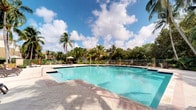 This outdoor amenity area centers around a large, resort-style swimming pool with clear turquoise water, designed to accommodate both leisurely relaxation and active swimming. The pool deck is finished with patterned, textured tile pavers in warm earth tones, providing a durable and visually appealing surface around the water's edge. Entry points are accessible via wide steps and stainless steel handrails, ensuring ease of access.

Surrounding the pool is an open-air deck furnished with lounge seating and at least one shaded umbrella station, offering comfort for sunbathing and socializing. A perimeter fence encloses the area, providing both security and defined boundaries for the space. Potted plants and tropical landscaping add decorative greenery along the fenceline.

The setting is framed by an abundance of mature palm trees and dense tropical foliage, creating a lush, private atmosphere reminiscent of a Florida resort environment. The open sky above allows for ample natural sunlight throughout the day. Neighboring rooftops visible in the background suggest this pool is part of a larger residential community, serving as a shared outdoor recreational amenity.