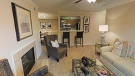 This single-floor residence offers a thoughtfully designed open-concept layout that seamlessly connects the living, dining, and kitchen areas. The living room serves as the heart of the home, featuring warm carpeted flooring, a classic fireplace with a tiled surround, and a ceiling fan overhead. Framed wall art and neutral-toned walls contribute to the space's inviting and comfortable ambiance. A woven accent chair and glass-top coffee table add stylish, functional touches to the seating area. The kitchen is accessible via a pass-through breakfast bar with raised countertop seating, making it ideal for casual dining and entertaining. Dark wood cabinetry, stainless steel appliances — including a microwave and refrigerator — and ample counter space give the kitchen a practical yet polished look. Track lighting illuminates the kitchen area efficiently. The dining area is visible beyond the living room, benefiting from the open sightlines of the floor plan and additional track lighting. Crown molding details add a refined architectural touch throughout the space, enhancing the overall sense of quality and care in this well-maintained single-floor home.