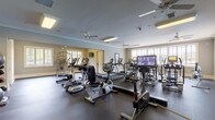 This fitness center offers a comprehensive workout environment designed to accommodate a variety of exercise routines. The main floor is generously sized, featuring durable rubber flooring throughout and multiple ceiling fans that help maintain a comfortable atmosphere during workouts. Large windows along the perimeter walls allow natural light to fill the space, creating an open and energizing environment.

The cardio area is prominently stocked with True-brand equipment, including multiple treadmills, elliptical trainers, rowing machines, and stationary bikes—one of which features a large integrated display screen. A dedicated section houses a rack of kettlebells and additional strength training equipment, providing options for resistance-based workouts. A separate, private alcove off the main floor contains a specialized body composition assessment station, offering a quiet space for fitness evaluations.

Crown molding and recessed lighting contribute a polished aesthetic to the space, while multiple ceiling fans are evenly distributed to ensure consistent air circulation. Motivational wall art adds character throughout. The layout is thoughtfully organized to allow users to move freely between equipment, making the fitness center both functional and welcoming for all levels of exercise experience.