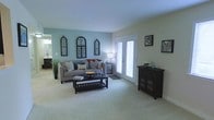 This single-floor space presents a welcoming living room that balances comfort and style. The room is painted in a soft, muted teal-green palette that creates a calm and airy atmosphere. Plush wall-to-wall carpeting in a light neutral tone runs throughout, adding warmth underfoot. A set of white French doors floods the space with natural light and provides access to the outdoors, contributing to the open and spacious feel of the room. A comfortable sectional sofa anchors the seating area, complemented by a dark wood coffee table with a lower shelf for additional storage. Decorative arched wall panels with an ornate, gothic-inspired design serve as a distinctive focal point on one wall, adding architectural character to the space. Framed artwork on the adjacent wall contributes further visual interest. A dark wood cabinet with louvered door panels provides functional storage while reinforcing the room's cohesive design aesthetic. A glimpse into an adjoining area, likely a kitchen, is visible through an open doorway, suggesting a connected and practical layout. Overall, the space offers a bright, well-appointed environment with thoughtful decorative details.