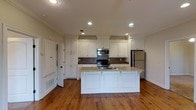 This single-floor space showcases a thoughtfully designed kitchen that serves as its centerpiece. The kitchen features classic white raised-panel cabinetry that extends to the ceiling, providing generous storage throughout. A decorative tile backsplash with a diamond pattern adds visual interest behind the range, complementing the stainless steel appliances, which include a built-in microwave, electric range, and a full-size refrigerator. The kitchen island is finished with elegant granite countertops and white wainscoting panels, offering additional workspace and a convenient sink. Recessed lighting illuminates the space evenly, enhancing the bright and clean aesthetic of the white cabinetry. Warm-toned hardwood floors run throughout, adding richness and character to the overall ambiance. Crown molding details along the ceiling lend a refined, traditional touch to the space. A doorway visible to the side suggests easy flow and connectivity to adjacent areas of the floor plan. The overall design strikes a balance between functional practicality and classic charm, making it a welcoming and highly usable space.