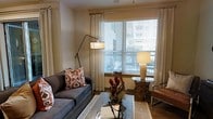 This single-floor space presents a thoughtfully designed interior that balances comfort and style. The living area serves as the heart of the home, showcasing warm neutral walls that complement the rich hardwood flooring beneath. A large window dressed with sheer floor-length curtains and horizontal blinds allows an abundance of natural light to fill the room, creating a bright and welcoming atmosphere while offering views of the surrounding exterior. The living area is furnished with a plush charcoal sofa accented by vibrant ikat-patterned throw pillows in warm rust and cream tones, adding a pop of texture and color to the space. An articulating arc floor lamp provides focused ambient lighting, while a ceramic table lamp atop a sculptural driftwood side table adds an organic, artistic touch. A mirrored coffee table introduces a modern element, reflecting light and contributing to the room's sense of openness. A leather accent chair rounds out the seating arrangement, lending a classic contrast to the softer furnishings. The overall ambiance is warm and inviting, with a carefully layered mix of natural textures, eclectic accents, and curated decor that gives the space a lived-in yet polished character.