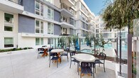 This amenity-rich courtyard space is set within a contemporary multi-story residential building, designed to offer a resort-like outdoor experience at the ground level. The centerpiece is a generously sized rectangular swimming pool with crystal-clear turquoise water, framed by clean tile edging and a broad surrounding deck. A dedicated sunbathing area lines one side of the pool, outfitted with a long row of white lounge chairs positioned to maximize relaxation and open-air enjoyment. Glass panel fencing surrounds the pool area, maintaining visibility while providing a sense of security and openness. Adjacent to the pool deck, a well-appointed outdoor lounge and dining terrace features multiple round tables paired with modern rope-woven chairs, ideal for social gatherings or casual outdoor dining. Mature trees and ornamental plantings are interspersed throughout the courtyard, introducing natural greenery against the crisp, modern architecture. The surrounding building facade is finished in contrasting gray and white tones, with large multi-pane windows and private balconies overlooking the courtyard on upper floors. The open-air design allows abundant natural light to fill the space throughout the day, creating a bright and welcoming atmosphere for residents.