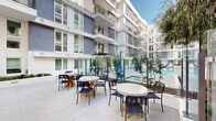This amenity-rich courtyard space is set within a contemporary multi-story residential building, designed to offer a resort-like outdoor experience at the ground level. The centerpiece is a generously sized rectangular swimming pool with crystal-clear turquoise water, framed by clean tile edging and a broad surrounding deck. A dedicated sunbathing area lines one side of the pool, outfitted with a long row of white lounge chairs positioned to maximize relaxation and open-air enjoyment. Glass panel fencing surrounds the pool area, maintaining visibility while providing a sense of security and openness. Adjacent to the pool deck, a well-appointed outdoor lounge and dining terrace features multiple round tables paired with modern rope-woven chairs, ideal for social gatherings or casual outdoor dining. Mature trees and ornamental plantings are interspersed throughout the courtyard, introducing natural greenery against the crisp, modern architecture. The surrounding building facade is finished in contrasting gray and white tones, with large multi-pane windows and private balconies overlooking the courtyard on upper floors. The open-air design allows abundant natural light to fill the space throughout the day, creating a bright and welcoming atmosphere for residents.