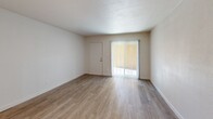 Located at 2912 East Indian School Road in Phoenix, Arizona, this single-floor unit offers a efficiently designed layout centered around a bright, open living room. Light wood laminate flooring runs throughout the space, contributing to a clean and cohesive aesthetic. The walls are finished in crisp white, enhancing the sense of openness and reflecting natural light that enters through a sliding glass door fitted with vertical blinds. The sliding door provides direct access to an outdoor deck area featuring wood plank decking and a stucco exterior wall, with lush greenery and palm trees visible in the surrounding landscape — a nod to the classic Phoenix environment.

The living area flows naturally toward a kitchen counter bar with granite stone countertops, providing a functional transition between the living and kitchen spaces. Recessed lighting and a ceiling-mounted HVAC vent ensure the kitchen area is well-lit and climate-controlled. In-unit laundry is conveniently integrated into the space, with a washer unit positioned near an electrical panel and an interior door. A textured ceiling adds subtle architectural character throughout. The overall design is clean, modern, and move-in ready, making efficient and practical use of the available floor area.