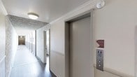 This single-floor space is accessed via a shared hallway that reflects a functional and well-maintained design. The corridor features dark hardwood flooring that runs the length of the passage, providing a warm contrast to the neutral-toned walls accented with textured stone or marble-patterned wall panels, adding a touch of visual interest to the common area. Crown molding along the ceiling contributes a classic architectural detail, lending the space a refined character. An elevator is conveniently located within the hallway, fitted with standard call buttons and surrounded by safety signage, including fire emergency instructions and a smoke-free environment notice — indicating a professionally managed building. Ceiling-mounted flush lighting illuminates the corridor evenly, while a smoke detector is visibly installed for safety compliance. The hallway extends toward additional doorways, suggesting multiple units or rooms accessible from this shared passage. Overall, the space presents a clean, orderly common area that balances practicality with modest architectural detailing, creating a functional environment suited to daily use.