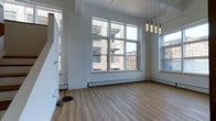 This single-floor space offers a clean, open layout defined by a crisp white interior and warm hardwood flooring that runs throughout. Large, multi-pane windows dominate the main living area, flooding the space with abundant natural light and framing views of the surrounding urban streetscape and neighboring brick buildings. The generous window area creates a strong visual connection to the city outside while enhancing the overall sense of openness.

A set of stylish pendant lights hangs from the ceiling, providing a modern accent that complements the otherwise minimalist aesthetic. Baseboard heating runs beneath the windows, ensuring comfort throughout the seasons. The clean white walls and light-toned floors give the space a fresh, airy atmosphere that lends itself well to a variety of interior arrangements.

Structural elements such as visible ceiling beams add a subtle industrial character to the space, referencing the building's architectural heritage while maintaining a contemporary feel. A staircase is visible along one wall, hinting at the vertical layout of the broader building. Overall, this space presents a well-lit, versatile environment with urban character, modern finishes, and a bright, welcoming ambiance throughout.