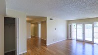 This single-floor space offers an open, airy layout highlighted by warm hardwood flooring that extends throughout the main living areas. The neutral beige walls provide a clean, versatile backdrop suitable for a variety of interior styles. A textured popcorn ceiling adds a classic touch overhead, while a centrally located HVAC vent ensures comfortable climate control throughout the space.

The main living area is generously proportioned, with a glass-paneled exterior door along one wall that allows natural light to pour into the room, enhancing the sense of openness. This door also provides a view of the greenery outside, connecting the interior to the outdoors.

A wide opening in the interior wall leads to a hallway, providing access to additional rooms beyond. The transition between spaces is smooth and unobstructed, supporting an easy flow throughout the layout. White baseboards frame the flooring cleanly, adding a crisp finish to each room.

Overall, the space presents a straightforward, functional design with a bright and neutral ambiance, making it a practical and adaptable living environment.