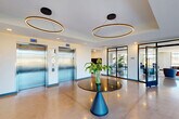 Located at 2310 Cochrane St in Detroit, Michigan, this contemporary ground-floor space offers a generously sized, open-plan layout spread across a single level. The design is distinctly modern, characterized by polished concrete flooring that reflects light throughout and crisp white walls that create a clean, professional atmosphere.

The main lobby serves as the centerpiece of the space, featuring two stainless steel elevators, a built-in mailbox wall, and a striking circular ring pendant light fixture suspended above a sleek pedestal table adorned with fresh tropical foliage. Floor-to-ceiling windows and glass-paneled entry doors flood the area with abundant natural light and provide clear views of the surrounding Detroit streetscape.

A connected lounge and social area is furnished with a mix of seating arrangements—including a sofa, accent chairs, and multiple dining-height tables with chairs—making it well-suited for both casual gatherings and informal meetings. A compact kitchenette with counter seating, a microwave, and cabinetry adds functional convenience to this communal zone.

Throughout the space, thoughtful touches such as potted tropical plants, curated artwork, and carefully selected furnishings contribute to a welcoming and polished ambiance. The open layout, generous glazing, and cohesive modern aesthetic make this a distinctive and well-appointed ground-floor destination.