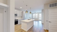 This single-floor residence showcases a thoughtfully designed open-concept layout that seamlessly connects the kitchen and living areas. The kitchen is a standout feature, outfitted with crisp white shaker cabinetry, light quartz countertops, and a full suite of stainless steel appliances, including a French door refrigerator with ice and water dispenser, a slide-in range, built-in microwave, and dishwasher. A generously sized center island with an undermount sink serves as the focal point, illuminated by a pair of striking brushed gold pendant lights. Light wood-style flooring flows throughout the main living spaces, complementing the neutral palette and bright, airy atmosphere. The living area benefits from abundant natural light through large windows and a glass door that opens to an outdoor balcony, complete with a ceiling fan overhead. The bedrooms offer plush carpeting underfoot, ceiling fans, and ample natural light, with one bedroom featuring a spacious walk-in closet equipped with wire shelving for organized storage. The bathroom is finished with a white vanity, quartz countertop, brushed gold fixtures, a large framed mirror, and a full tub-shower combination with tiled surround. Throughout the space, neutral tones and consistent gold-toned hardware tie each room together cohesively.