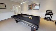 This single-level game room is designed with both style and function in mind, making it an inviting space for recreation and socializing. The centerpiece is a full-size billiard table finished in black felt, complemented by matching dark wood legs and trim that lend a sophisticated feel to the room. A distinctive curved arc pendant light fixture hangs overhead, providing focused illumination directly over the table, while recessed ceiling lights offer additional ambient lighting throughout the space.

The room features classic architectural detailing, including crown molding along the ceiling perimeter and a chair-rail wainscoting treatment along the walls, painted in a warm neutral tone. Neutral-toned carpet covers the floor, adding comfort underfoot. A set of tall black spectator chairs lines the perimeter, providing ample seating for observers. Windows fitted with white horizontal blinds allow natural light to filter into the room while maintaining privacy.

Framed abstract artwork adorns the walls, adding visual interest and a contemporary touch. A set of French doors with glass panels connects the space to an adjacent area, enhancing the open and accessible feel. Overall, the room balances a refined aesthetic with a relaxed, recreational atmosphere.