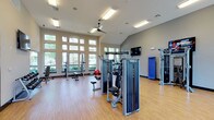 This single-level fitness center offers a thoughtfully designed workout environment suited for a range of exercise routines. The space is anchored by tall, multi-pane windows along one wall, flooding the interior with natural light and offering views of the surrounding greenery. Vaulted ceilings enhance the sense of openness, while ceiling fans and recessed fluorescent lighting ensure a comfortable and well-lit atmosphere throughout.

The flooring consists of warm hardwood-style material, providing a clean and durable surface suitable for both equipment use and open-floor exercises. The facility is outfitted with a selection of modern strength-training machines, including cable and weight systems, along with free weights and benches for versatile workouts. A wall-mounted flat-screen television is positioned for visibility from the main workout area, adding an entertainment option for users.

Additional equipment, including resistance and functional training apparatus, is arranged along the perimeter, maximizing usable floor space. The neutral color palette of the walls contributes to the bright, clean aesthetic of the space, making it an inviting and functional environment for fitness activities.