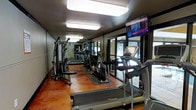 The Stonemill Village fitness center is a dedicated, single-floor workout facility designed to meet a variety of exercise needs. The gym is outfitted with a range of cardio equipment, including treadmills and an elliptical machine, complemented by a multi-station strength training unit and a stationary bike. The space is anchored by a warm, richly toned polished concrete floor that adds character and durability underfoot. Overhead, flush-mounted fluorescent lighting ensures the entire area is brightly and evenly illuminated, creating a clear and energizing workout environment. A wall-mounted flat-screen television provides entertainment and connectivity, keeping residents informed and engaged during their sessions. Large windows and glass doors line one wall, inviting an abundance of natural light and offering views of the surrounding outdoor area, enhancing the overall ambiance of the space. Strategically placed mirrors amplify the sense of openness and allow users to monitor their form during workouts. The layout is thoughtfully organized to allow comfortable movement between equipment stations. Overall, the fitness center presents a functional and well-maintained amenity that supports an active lifestyle for residents of Stonemill Village.