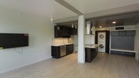 This single-floor space showcases a contemporary open-concept layout that seamlessly connects the living area and kitchen, creating a functional and visually cohesive environment. The kitchen is outfitted with dark, floor-to-ceiling cabinetry that provides generous storage, complemented by light-toned countertops that offer a striking contrast. Stainless steel appliances, including a refrigerator and dishwasher, add a polished, modern finish, while a wall-mounted range hood anchors the cooking area. Track lighting runs along the ceiling, providing focused illumination throughout the kitchen space. A structural column serves as a subtle architectural divider between the kitchen and an adjacent area that houses a stacked washer and dryer unit — a highly practical in-unit convenience. The light wood-toned flooring flows continuously throughout, lending warmth and cohesion to the overall aesthetic. Neutral gray walls keep the palette clean and versatile, while the open layout enhances the sense of spaciousness. The design balances modern industrial elements with everyday functionality, making it a well-appointed and efficient living space.