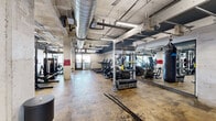 This well-equipped fitness center offers a generous single-floor layout with an industrial aesthetic that sets it apart from conventional gyms. Exposed concrete columns, polished concrete flooring, and an open ceiling with visible ductwork — including a prominent spiral ventilation duct — give the space a raw, urban character. Linear LED lighting runs throughout, ensuring consistent, bright illumination across all workout zones.

The cardio section lines one side of the floor and includes multiple treadmills, upright and recumbent bikes, stair climbers, and elliptical machines — all from Matrix, a commercial-grade equipment brand. Floor-to-ceiling mirrors along this wall expand the sense of space and allow users to monitor their form during workouts.

The strength training area is equally well-appointed, featuring heavy-duty power racks and squat stands loaded with Olympic barbells and weight plates, along with a variety of selectorized weight machines for targeted muscle training. Rubber mat flooring beneath the free weight stations provides safety and durability. A heavy punching bag is also present, adding a functional training dimension to the space.

Open floor areas provide room for stretching, bodyweight exercises, or functional fitness movements. Natural light filters in through windows near the entrance, complementing the overhead lighting. The overall ambiance is purposeful and performance-oriented, designed to support a wide range of fitness goals.