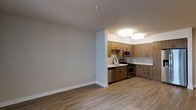 This single-floor space showcases a clean, contemporary design with an open-concept layout that connects the living area to a fully equipped kitchen. The living space features neutral gray walls and light hardwood-style flooring that extends throughout, creating a cohesive and airy feel.

The kitchen is thoughtfully designed with warm, medium-toned wood cabinetry spanning both upper and lower sections, offering generous storage. A classic subway tile backsplash adds texture and contrast, complementing the stainless steel appliances, which include a gas range, dishwasher, and over-the-range microwave. The countertops provide ample workspace, and the sink is positioned beneath upper cabinetry for practical use.

Overhead, flush-mount square light fixtures illuminate the kitchen efficiently, while recessed lighting in the living area maintains a clean ceiling profile. The neutral color palette of whites and grays throughout the space lends a modern, understated aesthetic. The open layout between the kitchen and living area enhances the sense of spaciousness, making the unit feel bright and functional.