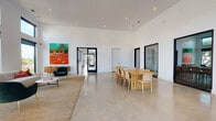 This contemporary single-floor space is defined by its striking architectural design and open, airy layout. Soaring ceilings rise dramatically throughout, amplifying the sense of volume and grandeur across the main areas. Polished concrete floors run seamlessly underfoot, lending a clean, industrial-modern aesthetic that ties the entire space together.

The main area is divided into two distinct yet connected zones. The lounge area features a comfortable neutral sofa accented with vibrant throw pillows, a sleek marble coffee table, and a bold emerald green accent chair — all anchored by a woven jute rug. A large, colorful contemporary painting of a tennis court scene serves as a striking focal point on the white wall, adding energy and personality to the space.

Adjacent to the lounge, a generously sized natural wood dining table with rattan-backed chairs accommodates a large group, making it well-suited for gatherings or collaborative use.

Floor-to-ceiling black-framed windows and glazed doors line the walls, flooding the interior with natural light and offering views of the surrounding landscape, including mature trees and outdoor structures. Multiple doorways connect to additional rooms and exterior spaces, enhancing the flow and accessibility of the layout. A reception-style counter near the entrance adds a functional, professional touch to the overall design.