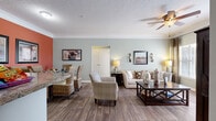Located at 150 Carrington Park Dr in Gainesville, Georgia, this single-floor residence offers a thoughtfully designed layout that balances comfort and functionality throughout. The open-concept living and dining area serves as the heart of the home, featuring durable wood-look flooring, crown molding, and a textured ceiling. Soft sage-green walls in the living room contrast with a warm terracotta accent wall in the dining area, creating a welcoming and visually dynamic atmosphere. A ceiling fan overhead and wall sconce lighting add both practicality and character. The dining area accommodates a full table and wicker chairs, while the living room provides ample space for relaxed seating around a classic wood coffee table. The galley-style kitchen is equipped with honey-toned wood cabinetry, granite-look countertops, and white appliances including a dishwasher and range, with wood-look flooring continuing the cohesive design. The bedroom offers plush carpeting, a ceiling fan, and natural light from a window, with direct access to a private en suite bathroom featuring a wide vanity and tub-shower combination. A spacious walk-in closet with wire shelving provides generous storage. A second full bathroom with wood-look flooring, a large mirror, and a tub-shower rounds out this comfortable and well-organized home.