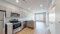 This contemporary single-floor unit is designed with a clean, modern aesthetic throughout. The open-concept layout seamlessly connects the kitchen and living area, creating a bright and functional space ideal for everyday living. The kitchen is well-appointed with grey shaker-style cabinetry, light granite-look countertops, and a full suite of stainless steel appliances including a refrigerator, electric range, over-the-range microwave, and dishwasher. An L-shaped peninsula with an integrated sink defines the kitchen boundary and provides additional prep and seating space. Recessed lighting illuminates the area efficiently, complementing the natural light that filters in through large windows dressed with horizontal blinds. Consistent light-toned hardwood flooring flows throughout the entire unit, lending a cohesive and airy feel. The living area is generously proportioned and open, offering flexibility for various furniture arrangements. The bedroom features a ceiling fan, a window with an outdoor view, and ample wall space. The bathroom is neatly finished with a grey vanity topped with a granite-look countertop, a toilet, and a full tub/shower combination unit. Neutral grey walls and clean white trim tie the design together, creating a polished and move-in-ready environment.