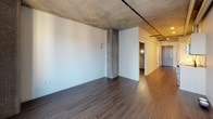 This single-floor urban residence showcases a compelling blend of industrial and modern design elements throughout. The open living area immediately draws attention with its raw exposed concrete ceiling and visible silver ductwork running overhead, lending the space a distinctly loft-inspired character. Dark hardwood flooring flows continuously throughout, grounding the airy, light-toned walls and creating a warm contrast against the industrial ceiling.

Large windows in the main living area flood the space with natural light and frame views of the surrounding urban cityscape, offering a vivid connection to the metropolitan environment outside. Exposed concrete structural columns punctuate the open layout, reinforcing the architectural identity of the space.

The kitchen is well-appointed with light wood cabinetry, white quartz countertops, a classic white subway tile backsplash, and a full suite of stainless steel appliances including a refrigerator, electric range, built-in microwave, and dishwasher — combining style with functional efficiency.

The bathroom features a clean, contemporary design with a light wood vanity, white countertop, a full tub-shower combination, and neutral tile flooring, creating a calm and practical retreat.

Overall, the space delivers a cohesive industrial-modern aesthetic with an open, airy feel enhanced by city views and quality finishes throughout.