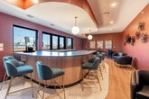 Located at 425 SW 11th St in Des Moines, Iowa, this sophisticated rooftop lounge and kitchen space offers a compelling blend of contemporary design and urban ambiance. The interior centers around a striking curved bar with a sleek dark countertop and warm wood paneling, surrounded by teal velvet barstools with gold-toned legs set atop a hexagonal tile inlay. Globe pendant lights hang from a sculptural curved ceiling detail finished in rich copper tones, casting a warm glow over the space. Deep mauve and terracotta walls are adorned with unique sculptural wall art, contributing to the space's upscale, curated aesthetic. Light hardwood flooring runs throughout, complementing the eclectic mix of seating that includes leather lounge chairs and bistro-style tables. The kitchen area features stone countertops, under-counter refrigeration units, and ample prep space, making it well-suited for entertaining. Large windows frame views of the Des Moines skyline, filling the interior with natural light. The expansive outdoor terrace is finished with large-format tile, warm wood privacy panels, and strung Edison bulb lighting, offering unobstructed panoramic views of the downtown skyline — an ideal setting for gatherings day or night.