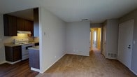 This single-floor residence offers a practical and straightforward layout designed for comfortable everyday living. The open-concept living area is finished with wall-to-wall carpeting in a warm neutral tone, creating a cozy and inviting atmosphere. Smooth, light-gray walls and a textured popcorn ceiling carry throughout the main living space, contributing to a clean and consistent aesthetic.

Adjacent to the living area is the kitchen, which is partially separated by a half-wall partition. The kitchen features dark wood cabinetry, a countertop with a built-in range, and a tile backsplash, offering functional workspace and ample storage. The transition from hardwood-style flooring in the kitchen to carpet in the living area subtly defines the two zones without the need for a full partition.

A hallway extends from the living area toward the rear of the unit, providing access to additional rooms. Doors along the hallway suggest separate spaces such as bedrooms or a bathroom. Natural light filters through what appears to be a window at the end of the hallway, brightening the passage and adding a sense of depth to the layout.

Overall, this space presents a no-frills, functional design with consistent finishes throughout, well-suited for those seeking a straightforward and practical living environment.