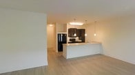 This single-floor space showcases an open-concept design that integrates the kitchen and living area into one cohesive, flowing environment. The light hardwood-style flooring runs throughout, lending a warm and cohesive aesthetic across the space.

The kitchen is a standout feature, appointed with rich dark cabinetry that provides a striking contrast against the light-toned countertops and white walls. A peninsula-style counter extends outward, offering additional workspace and serving as a natural divider between the kitchen and living area. Three pendant lights hang above the peninsula, providing both functional task lighting and a modern decorative element. A flush-mounted ceiling fixture above the main cooking area adds further illumination to the workspace. Black appliances, including a refrigerator and range, complement the dark cabinetry for a consistent, contemporary look.

The living area benefits from the open sightlines created by the layout, making the space feel generously sized. Soft, ambient lighting throughout contributes to a warm and inviting atmosphere. The overall design balances functionality with modern aesthetics, creating a practical yet stylish environment suited to everyday living.