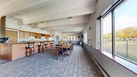 Located in Caledonia, Wisconsin, this single-floor clubhouse offers a well-appointed communal space designed for gathering and entertaining. The open-plan layout is anchored by exposed beam ceilings that lend architectural character and a sense of volume to the interior. Large windows line the walls, flooding the space with natural light and providing pleasant views of the surrounding outdoor landscape.

The main gathering area features patterned carpet flooring and accommodates multiple dining tables and chairs, making it well-suited for group events and social functions. A standout design element is the diagonally planked wood accent wall, which adds warmth and texture to the dining area, complemented by track lighting overhead.

The kitchen area is fully equipped with wood cabinetry, a granite-topped island with bar-height seating, a refrigerator, microwave, range, and sink — providing everything needed to support catered events or casual gatherings. Pendant lighting above the island enhances both function and ambiance. A coffee station is also present near the kitchen counter area.

The overall atmosphere is warm and welcoming, balancing functional amenities with comfortable, community-oriented design. The space is well-lit, open, and thoughtfully arranged to serve the needs of residents and groups alike.