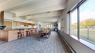 Located in Caledonia, Wisconsin, this single-floor clubhouse offers a well-appointed communal space designed for gathering and entertaining. The open-plan layout is anchored by exposed beam ceilings that lend architectural character and a sense of volume to the interior. Large windows line the walls, flooding the space with natural light and providing pleasant views of the surrounding outdoor landscape.

The main gathering area features patterned carpet flooring and accommodates multiple dining tables and chairs, making it well-suited for group events and social functions. A standout design element is the diagonally planked wood accent wall, which adds warmth and texture to the dining area, complemented by track lighting overhead.

The kitchen area is fully equipped with wood cabinetry, a granite-topped island with bar-height seating, a refrigerator, microwave, range, and sink — providing everything needed to support catered events or casual gatherings. Pendant lighting above the island enhances both function and ambiance. A coffee station is also present near the kitchen counter area.

The overall atmosphere is warm and welcoming, balancing functional amenities with comfortable, community-oriented design. The space is well-lit, open, and thoughtfully arranged to serve the needs of residents and groups alike.