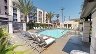 This outdoor amenity space offers a well-appointed resort-style experience designed for relaxation and recreation. At its center is a generously sized swimming pool with clear, blue water, surrounded by a spacious concrete sundeck lined with modern lounge chairs and shaded by market umbrellas — ideal for soaking up the sun or unwinding in comfort. Tall palm trees are thoughtfully integrated throughout the pool area, adding a Southern California ambiance and natural shade.

Adjacent to the pool, a dedicated outdoor kitchen area provides a stainless steel built-in BBQ grill set into a stone countertop, sheltered beneath a corrugated metal pergola roof supported by masonry columns. A dining table and chairs complete the space, making it well-suited for outdoor gatherings and al fresco dining. Lush greenery and manicured shrubs line the perimeter walls, adding privacy and a vibrant touch of nature to the space.

The entire area is enclosed by masonry block and iron fencing, providing security while maintaining an open, airy feel. Pool rules and capacity signage are posted on-site. The surrounding multi-story building with balconies frames the courtyard, creating a cohesive and well-designed community outdoor environment under open blue skies.