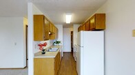 This single-floor residence offers a practical and well-organized layout designed for comfortable everyday living. The heart of the home is a classic galley-style kitchen, efficiently arranged to maximize both storage and workflow. Warm honey-toned wood cabinetry lines both sides of the kitchen, providing generous storage space above and below the countertops. The light-colored countertops offer ample workspace, complemented by a double-basin sink positioned conveniently along one wall. White appliances, including a full-size refrigerator and dishwasher, contribute to the kitchen's clean and cohesive aesthetic. A flush-mounted ceiling light fixture brightens the space, while a textured ceiling adds a touch of classic character overhead. Hardwood-style flooring runs the length of the kitchen, offering both durability and a warm, inviting feel. The kitchen opens toward a hallway that connects to the remaining areas of the residence, suggesting a thoughtful and accessible floor plan. Small decorative touches, such as potted plants and floral accents, enhance the welcoming atmosphere of the space. Overall, this residence presents a tidy, functional environment well-suited for practical, day-to-day living.