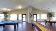 This single-story game room is designed for recreation and entertainment, showcasing a spacious, open layout enhanced by striking vaulted ceilings that add a sense of grandeur to the space. The soaring roofline draws the eye upward, creating an airy atmosphere throughout the room.

The hardwood flooring runs the length of the space, providing a warm, polished foundation that complements the room's overall aesthetic. A pool table anchors one side of the room, while a full-length shuffleboard table stretches along the opposite side, offering diverse entertainment options. Sports-themed wall decals add a playful, decorative touch to the central wall, reinforcing the room's recreational character.

Natural light filters in generously through multiple windows fitted with horizontal blinds, illuminating the space from several angles. Track lighting mounted on the ceiling provides additional, adjustable illumination for evening use.

The textured ceiling adds subtle visual interest overhead, while the neutral wall tones keep the space feeling open and inviting. Access to adjacent outdoor areas is provided through glass-paneled doors, further enhancing the connection between the interior and exterior environments.