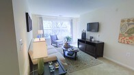 This single-floor space offers a warm and inviting atmosphere, thoughtfully designed for both comfort and functionality. The living area serves as the heart of the home, anchored by a plush sectional sofa and complemented by a patterned accent chair that adds a touch of personality to the room. A dark wood media console sits opposite the seating area, providing a clean and organized home for entertainment needs, including a wall-mounted flat-screen television.

Natural light flows generously into the space through a large window dressed with elegant curtains, offering views of mature trees and lush greenery outside. This connection to the outdoors enhances the room's airy and open feel. The neutral color palette of soft whites, beiges, and warm browns creates a calming ambiance throughout.

An oval coffee table at the center of the living area is styled with decorative accents, while a patterned area rug grounds the seating arrangement and adds visual interest to the light carpeted flooring. Tasteful lamp fixtures and curated décor pieces complete the refined aesthetic. The overall design strikes a harmonious balance between livability and style, making this a welcoming and well-presented space.