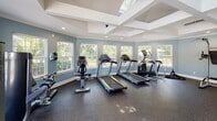 Located at 1904 US-80 in Garden City, Georgia, this single-story fitness center offers a comprehensive workout environment designed for both cardio and strength training. The space is defined by its distinctive coffered ceiling with skylights, which floods the interior with natural light and contributes to an open, airy atmosphere. Soft blue-gray walls and crisp white crown molding and ceiling details give the facility a clean, polished aesthetic throughout.

The cardio area is generously equipped with multiple treadmills, elliptical trainers, and stationary and recumbent bikes, all positioned along a perimeter of large windows that offer outdoor views of the surrounding greenery and pool area, creating an inviting environment for extended workouts. Durable rubber flooring throughout the facility provides both safety and comfort underfoot.

The strength training section includes a multi-function cable machine, a power tower, a weight bench, a Matrix selectorized machine, and a full rack of dumbbells. Stability and exercise balls are mounted on the wall for convenient access. Together, these areas provide users with a well-rounded, functional fitness facility suited to a wide range of exercise routines.