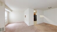 This single-floor unit offers three bedrooms and one bathroom within a efficiently organized layout. The living area is the most prominent shared space, featuring wall-to-wall carpeting in a neutral tone and a large sliding glass door fitted with vertical blinds, allowing natural light to fill the room and providing outdoor access. A secondary window on an adjacent wall further brightens the space. The living area connects to a partitioned wall that separates it from the kitchen, which features light wood-style flooring, warm-toned wood cabinetry, a black refrigerator, and a pass-through countertop ledge that opens toward the main living space.

The three bedrooms are carpeted and finished in neutral white and beige tones, each with standard windows that admit natural light. The primary bedroom offers a generous footprint among the three. The single bathroom includes a full-sized soaking tub with a white surround enclosure and chrome fixtures. Throughout the unit, the consistent use of light wall colors, neutral carpeting, and simple finishes creates a clean, functional ambiance suited for everyday living.