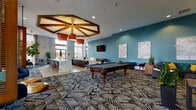 Located along El Camino Real in Santa Clara, California, this single-floor amenity clubhouse is thoughtfully designed to offer a dynamic social and recreational environment. The space is anchored by a generously sized open-plan lounge area that seamlessly integrates multiple activity zones under soaring ceilings that flood the interior with natural light through floor-to-ceiling windows dressed with sheer linen drapes.

A standout architectural detail is the geometric coffered ceiling feature crafted from warm wood beams, accented by a pair of cylindrical pendant lights, which frames a central games area complete with a classic billiards table. A dedicated shuffleboard corner, highlighted by a wall-mounted flat-screen TV and rich teal accent walls, provides additional entertainment options.

Comfortable lounge seating areas are distributed throughout, styled with bold teal and yellow accent pillows against a backdrop of curated abstract wall art and lush indoor plants. A sleek wet bar with marble-topped counters and bar seating anchors one end of the space, ideal for social gatherings. Double French doors open directly to an outdoor terrace, further enhancing the indoor-outdoor connectivity. The patterned carpet, contemporary furnishings, and warm wood tones throughout contribute to a cohesive, modern aesthetic that balances sophistication with approachable comfort.