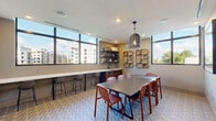 Located at 2850 Southwest 71st Avenue in Miami, FL, this well-appointed dining room offers a thoughtfully designed space ideal for communal gatherings and shared meals. The room is anchored by a large rectangular dining table surrounded by warm terracotta-toned chairs, providing ample seating for a group. A striking woven pendant light hangs overhead, adding a bohemian touch to the modern interior.

Floor-to-ceiling black-framed windows line one wall, flooding the space with natural light and offering views of the surrounding Miami skyline. Beneath the windows, a continuous white countertop serves as a casual bar-height seating area, complemented by sleek bar stools — perfect for individual use or socializing.

A built-in sink and dark wood cabinetry anchor one corner, while open shelving above displays decorative items and supplies. The opposite wall is adorned with a curated arrangement of woven rattan wall art, adding texture and visual warmth. A wall-mounted flat-screen television is also present, enhancing the room's versatility.

The patterned carpet in cool blue tones grounds the space with a contemporary flair, while recessed lighting and textured accent walls complete the refined, modern aesthetic. Framed artwork and smart home controls contribute to the polished, functional character of this inviting dining space.