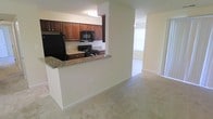 This single-floor residence offers a thoughtfully designed open-concept layout that promotes a sense of spaciousness and easy movement between living areas. The kitchen is a standout feature, showcasing rich dark wood cabinetry that provides ample storage, granite countertops with warm tonal variation, and black appliances including a refrigerator, gas range, and built-in microwave. A raised breakfast bar serves as a natural divider between the kitchen and the adjacent living space, making it ideal for casual dining or entertaining. The flooring transitions from the kitchen into soft, neutral-toned carpeting that extends through the main living area, adding warmth and comfort underfoot. The living area benefits from natural light filtering in through a window, contributing to a bright and welcoming ambiance. Neutral wall tones throughout the space provide a clean, versatile backdrop that complements a variety of décor styles. A ceiling fan is visible in the adjoining area, offering both practical comfort and a subtle architectural detail. The overall design is functional and cohesive, with a layout that maximizes usability while maintaining an open, airy feel throughout the single floor.