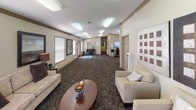 This single-floor space functions as a well-appointed community or leasing center, thoughtfully designed to welcome visitors with a warm, professional ambiance. The layout is open and elongated, allowing natural light to filter in through multiple blinded windows that frame views of surrounding greenery, contributing to an inviting atmosphere throughout.

The main lounge area is furnished with cream-upholstered armchairs and a sofa arranged around a rich, dark wood oval coffee table. Large-scale geometric wall art in contrasting tones serves as a striking focal point, while tall indoor palms add organic warmth to the corners of the room. Patterned, multi-tone carpeting runs throughout, grounding the neutral beige walls accented by classic crown molding.

Deeper into the space, a central hub features a wall-mounted flat-screen television, a water cooler, and a coffee station set atop a wooden cabinet — offering practical amenities for guests. An adjacent credenza area with decorative accessories and a mirror adds further character. A hallway leads to additional rooms and office areas visible in the background. Textured ceilings with flush lighting complete the space's clean, functional, yet comfortable aesthetic.