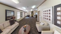 This single-floor space functions as a well-appointed community or leasing center, thoughtfully designed to welcome visitors with a warm, professional ambiance. The layout is open and elongated, allowing natural light to filter in through multiple blinded windows that frame views of surrounding greenery, contributing to an inviting atmosphere throughout.

The main lounge area is furnished with cream-upholstered armchairs and a sofa arranged around a rich, dark wood oval coffee table. Large-scale geometric wall art in contrasting tones serves as a striking focal point, while tall indoor palms add organic warmth to the corners of the room. Patterned, multi-tone carpeting runs throughout, grounding the neutral beige walls accented by classic crown molding.

Deeper into the space, a central hub features a wall-mounted flat-screen television, a water cooler, and a coffee station set atop a wooden cabinet — offering practical amenities for guests. An adjacent credenza area with decorative accessories and a mirror adds further character. A hallway leads to additional rooms and office areas visible in the background. Textured ceilings with flush lighting complete the space's clean, functional, yet comfortable aesthetic.