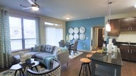 This single-floor residence showcases a thoughtfully designed open-concept layout that connects the living, dining, and kitchen areas into one cohesive and functional space. The living area is anchored by a tufted gray sectional sofa, complemented by patterned area rugs and a ceiling fan that adds both style and practicality. Large windows dressed with vertical blinds and decorative curtains allow generous natural light to fill the room, creating a bright and welcoming atmosphere.

A striking teal accent wall serves as the focal point of the dining area, adorned with a set of decorative circular wall pieces that add an artistic touch. The dining space comfortably accommodates a table and chairs, positioned atop a geometric-patterned rug. Dark hardwood-style flooring runs throughout, grounding the space with warmth and continuity.

An arched passageway leads toward what appears to be additional rooms, hinting at a well-connected floor plan. The kitchen, partially visible from the main living area, features dark cabinetry and is accessible via a breakfast bar with wooden bar stools, making it ideal for casual dining and entertaining.