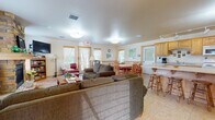 This single-story space is designed to accommodate group gatherings with a thoughtfully arranged, open floor plan. The main common area seamlessly connects a generously sized living room, multiple dining areas, and a fully equipped kitchen, creating a cohesive and functional environment.

The living room features a striking corner stone fireplace as its focal point, complemented by comfortable upholstered sofas, a red leather armchair with ottoman, a wall-mounted flat-screen TV, and a bookshelf stocked with reading materials. Large windows with blinds allow ample natural light throughout.

The kitchen is outfitted with light wood cabinetry, white appliances including a refrigerator, stove, and microwave, and a central island with bar stool seating. Track lighting illuminates the workspace efficiently. Adjacent dining areas offer multiple round and rectangular wooden tables with seating for larger groups, lit by pendant and track lighting and accented by bold red wainscoting in one section.

The accessible bathroom includes grab bars, a wide-clearance vanity, and tile flooring, meeting ADA standards. Throughout the space, light tile flooring, neutral walls with warm wood trim, and abundant windows create a bright, practical, and welcoming atmosphere suitable for community or group use.