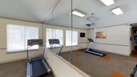 This fitness center is a thoughtfully designed single-floor exercise space that combines functionality with a bright, open atmosphere. The room features vaulted ceilings fitted with a ceiling fan, promoting air circulation during workouts, and is illuminated by a combination of recessed lighting and abundant natural light streaming through multiple large windows dressed with white blinds. The flooring consists of durable carpet with distinctive orange accent patches, adding a pop of color to the neutral palette of cream-toned walls. A full-length glass partition and wall-mounted mirrors visually expand the space while allowing members to monitor their form. Cardio equipment, including multiple treadmills, is positioned to take advantage of the natural light and open layout. A wall-mounted flat-screen television provides entertainment during workouts. Decorative artwork adorns the walls, contributing to a welcoming and motivating environment. The overall ambiance is clean, airy, and well-maintained, offering a comfortable setting for residents or members to pursue their fitness goals.