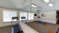 This fitness center is a thoughtfully designed single-floor exercise space that combines functionality with a bright, open atmosphere. The room features vaulted ceilings fitted with a ceiling fan, promoting air circulation during workouts, and is illuminated by a combination of recessed lighting and abundant natural light streaming through multiple large windows dressed with white blinds. The flooring consists of durable carpet with distinctive orange accent patches, adding a pop of color to the neutral palette of cream-toned walls. A full-length glass partition and wall-mounted mirrors visually expand the space while allowing members to monitor their form. Cardio equipment, including multiple treadmills, is positioned to take advantage of the natural light and open layout. A wall-mounted flat-screen television provides entertainment during workouts. Decorative artwork adorns the walls, contributing to a welcoming and motivating environment. The overall ambiance is clean, airy, and well-maintained, offering a comfortable setting for residents or members to pursue their fitness goals.