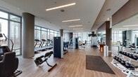 Located at 1201 N La Salle Drive in Chicago's Gold Coast neighborhood, this well-appointed fitness center offers a comprehensive, modern workout facility across a single open floor. Floor-to-ceiling windows line the perimeter, flooding the space with abundant natural light and providing panoramic views of the Chicago skyline and surrounding cityscape.

The facility is organized into distinct training zones, each designed for a specific type of exercise. The cardio area features a row of treadmills, elliptical machines, and stationary bikes, including a Peloton cycle, all positioned to take advantage of the expansive city views. A dedicated functional training section includes TRX suspension trainers, stability balls, battle ropes, and balance equipment, accompanied by a wall-mounted television for entertainment.

The strength training zone is fully equipped with a multi-station cable machine, a comprehensive selection of weight machines, and a dedicated free weights area outfitted with a full rack of dumbbells and kettlebells of varying sizes. Large mirrors along the walls enhance the sense of space and support proper form during workouts. A separate Pilates area houses a reformer and specialty equipment for low-impact training. The space features warm hardwood-style flooring throughout, a bold geometric accent wall in the free weights section, and elegant wood-panel entry doors, creating a polished and motivating environment.