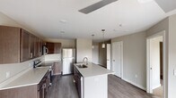 This single-floor residence is designed with a clean, contemporary aesthetic and a functional open-concept layout. The kitchen serves as the centerpiece of the living area, showcasing rich dark wood cabinetry paired with crisp light countertops for a striking contrast. A large center island with an integrated sink provides generous workspace and a casual gathering point, while three pendant lights hang overhead to create focused, stylish illumination. Stainless steel appliances — including a refrigerator, range, and built-in microwave — complete the well-equipped kitchen setup. The flooring throughout the main living area consists of durable wood-look planks in a warm neutral tone, lending the space a cohesive and modern feel. The open layout allows the kitchen to flow naturally into the adjacent living area, which benefits from a spacious and airy atmosphere with neutral wall tones. The bedroom is a comfortable and well-lit retreat featuring plush carpeting, a ceiling fan, and a generously sized window that invites natural light and outdoor views. The bathroom adjacent to the bedroom includes a dark wood vanity with a light countertop, a framed mirror with overhead lighting, and tile flooring, maintaining the consistent design language found throughout the residence.