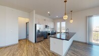 This thoughtfully designed single-floor residence showcases a contemporary aesthetic with high-quality finishes throughout. The open-concept kitchen is a standout feature, centered around a generous island with dark granite countertops, an undermount sink, and elegant gold pendant lighting above. Two-tone cabinetry pairs light wood upper cabinets with slate-blue lower cabinets, accented by brushed gold hardware. A white subway tile backsplash and a full suite of stainless steel appliances — including a French door refrigerator, range, over-the-range microwave, and dishwasher — complete the space. Durable wood-look flooring flows seamlessly through the kitchen and into the expansive open living area, which is bathed in natural light from large windows and features a ceiling fan overhead. The bedroom offers plush carpeting, dual windows with outdoor views, and a ceiling fan. A spacious walk-in closet provides ample built-in shelving and hanging storage. The primary bathroom impresses with slate-blue vanity cabinetry, granite countertops, wood-look flooring, and a soaking tub framed by marble-style tile surround. A secondary bathroom mirrors the same refined design with matching vanity and a tub-shower combination with marble-style tile walls.