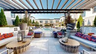 Located in the SouthPark neighborhood of Charlotte, NC, this resort-style outdoor amenity space offers a refined and thoughtfully designed pool courtyard that evokes a luxury retreat atmosphere. At its center sits a generously sized swimming pool with crystal-clear blue water, complemented by a shallow wading shelf with sculptural white in-pool lounge chairs — perfect for relaxing in the sun. Surrounding the pool, large-format light stone tile decking provides an expansive and clean foundation throughout the space. A striking wood-beam pergola anchors one end of the courtyard, sheltering a stylishly furnished lounge area with wicker seating, accent pillows, stone-clad fireplaces, and lantern lighting — ideal for social gatherings in any season. On the opposite side of the pool, rows of navy blue chaise lounges with coordinating side tables offer ample sunbathing space. Mature Italian cypress trees, potted greenery, and climbing ivy add layers of lush landscaping that soften the architecture and provide privacy. The surrounding multi-story building features a clean contemporary facade of white stucco, brick accents, and large-pane windows. The open sky above bathes the entire space in abundant natural light, creating a bright and inviting outdoor environment year-round.